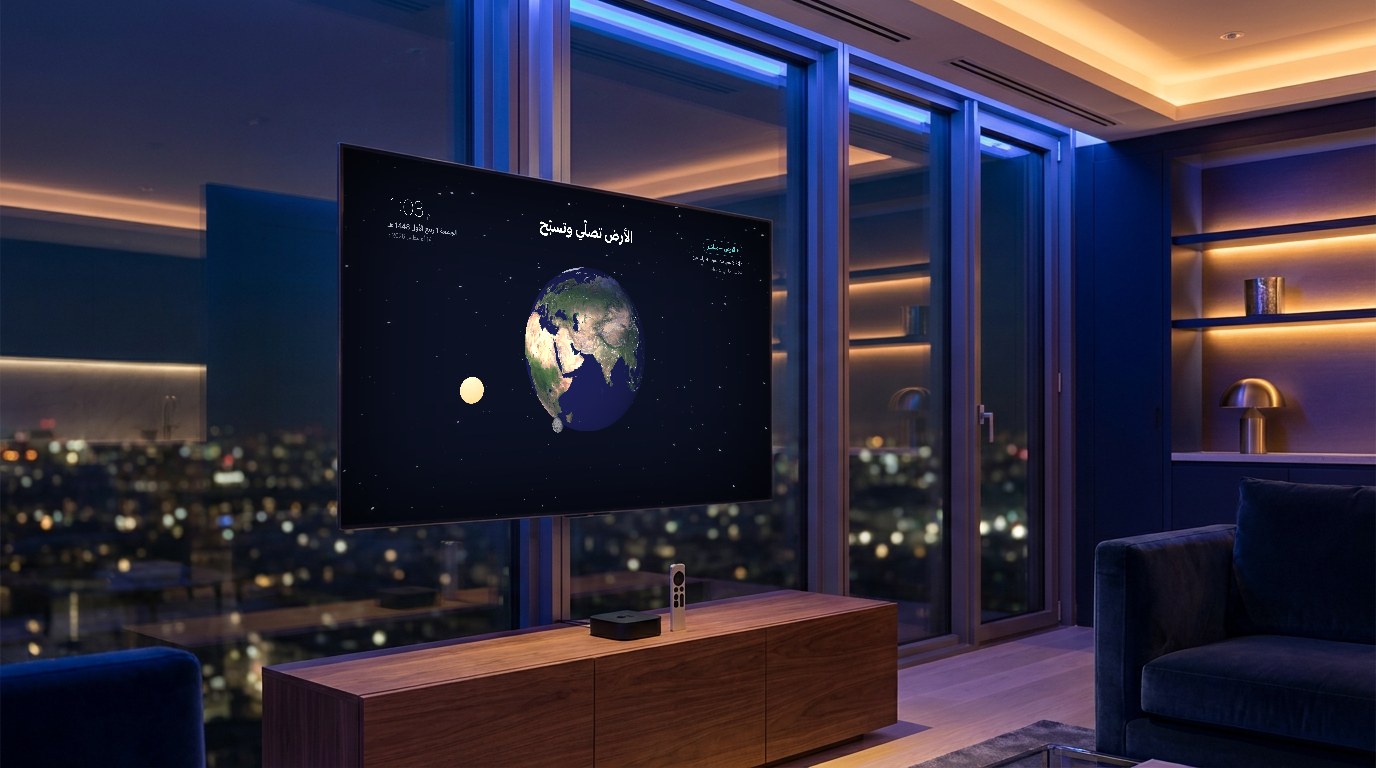 الأرض تصلّي وتسبّح على تلفازٍ في صالة برجٍ ليلاً — وجهاز Apple TV وريموته على الكونسول